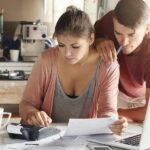 Two spouses sit at their kitchen table in front of their laptop, calculator, and paperwork to locate key tax savings and deductions in their finances.