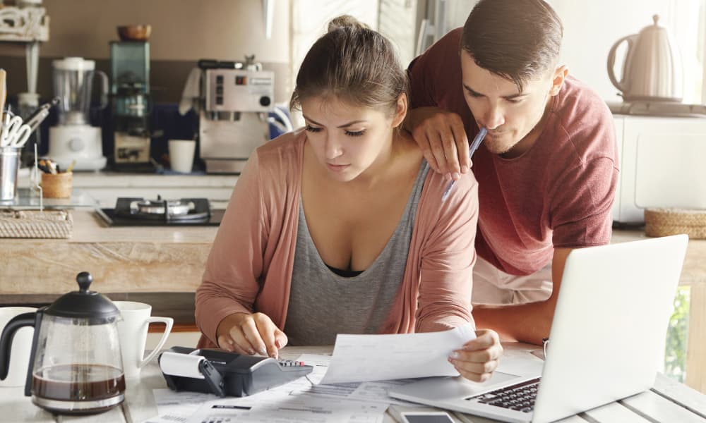 Two spouses sit at their kitchen table in front of their laptop, calculator, and paperwork to locate key tax savings and deductions in their finances.