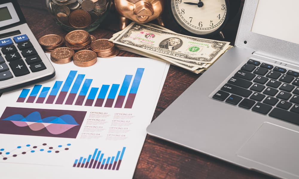 A paper with diagrams sits next to money and a laptop on a desk, illustrating the concept of saving money and investing.