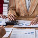 About 3 people are seen strategically tax planning by analyzing graphs and charts on a wooden table, using calculators to crunch numbers.