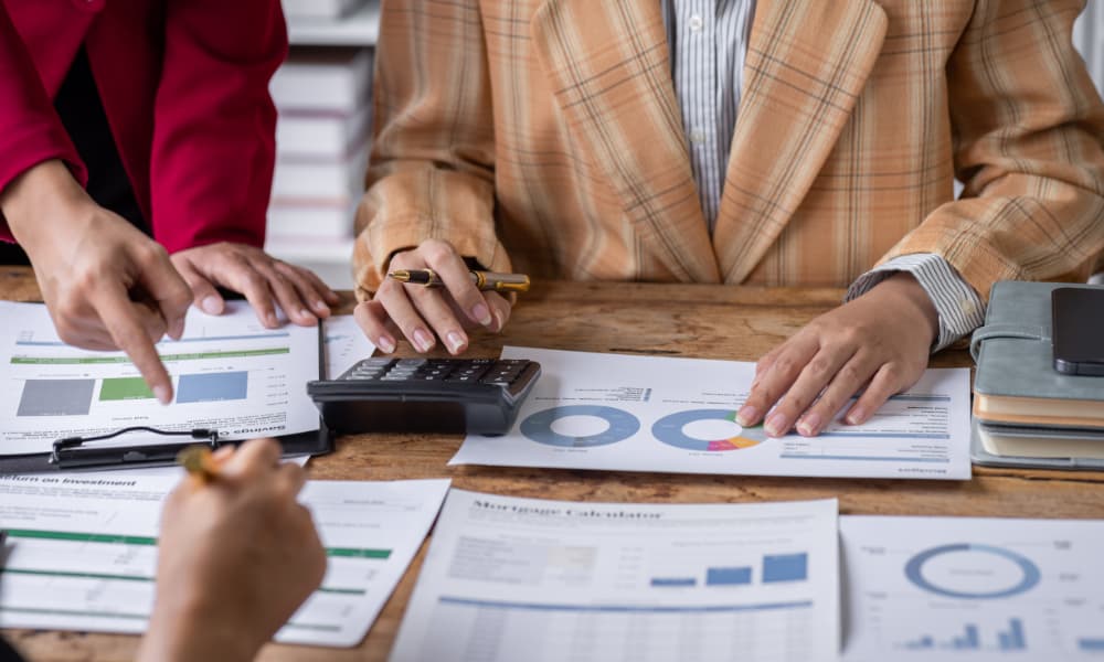 About 3 people are seen strategically tax planning by analyzing graphs and charts on a wooden table, using calculators to crunch numbers.