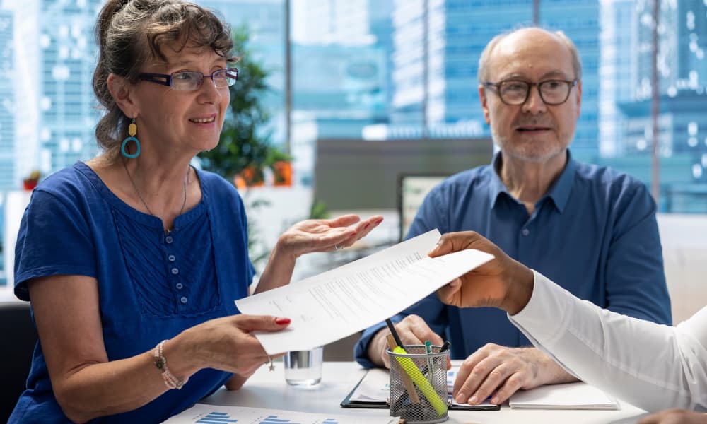 An elderly couple consults a financial advisor at a desk about making changes to their Medicare coverage.