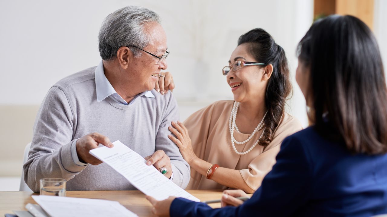 An elderly couple smiling while discussing estate planning documents with a financial advisor in an office setting.