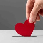 Close-up of a hand placing a red paper heart into a donation box sign of charitable contributions
